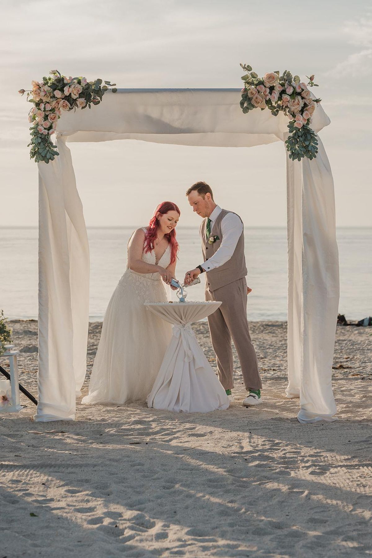 A bride and groom share a sand ceremony beneath an ivory floral arch on a Gulf Coast beach